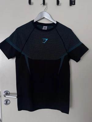 Svart och blå Gymshark t-shirt - Snygg svart t-shirt från Gymshark med blått mönster upptill och logga på bröstet. T-shirten har korta ärmar och är tillverkad i ett lätt och stretchigt material som passar perfekt till träning. Sportig design med coola detaljer. Aldrig använt den! Priset kan diskuteras
