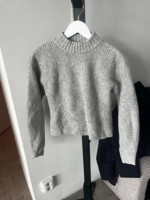Grå ribbstickad tröja från H&M - Säljer en grå ribbstickad tröja från H&M med hög krage och långa ärmar. Tröjan är i mjukt material och har en kortare passform som är perfekt att matcha med jeans eller kjol. Enkel och stilren design som passar till många olika outfits.