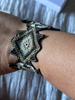 Mönstrat pärlarmband från Hipanema - Snyggt armband från Hipanema med pärlor i svart, beige och metallictoner. Armbandet har ett brett band med geometriskt mönster och silverfärgad magnetlås med Hipanema-logga. Perfekt för dig som gillar unika accessoarer.