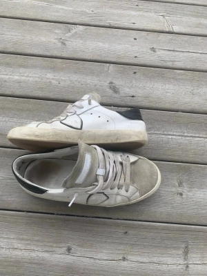Vita och grå sneakers med svart detalj - Snygga sneakers i vitt och grått med svart detalj på hälen. Ovandelen är i mocka och textil, med vita skosnören och perforerade sidor för extra stil. Klassisk låg modell som passar till det mesta. Perfekta för dig som gillar en clean och avslappnad look.