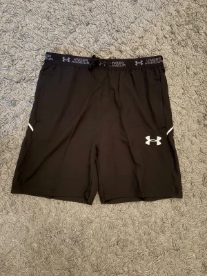 Under Armour shorts - Svarta träningsshorts från Under Armour med logga på benet och snörning i midjan. Shortsens material är lätt, perfekt för sport och aktivitet. Elastisk midja med vit text och diskreta vita detaljer på sidorna. M passar L