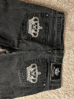 Svarta jeans med kronbrodyr - Säljer ett par svarta jeans från victoria Beckham med raka ben och stora fickor bak. På fickorna finns en snygg vit broderad krona som ger en unik look. Jeansen är i denim och har en klassisk passform med knapp och dragkedja.
