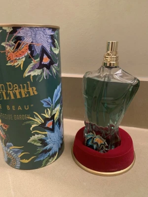 Jean Paul Gaultier Le Beau Paradise Garden - Parfymflaska formad som en manlig torso i transparent glas med turkosgrön vätska och guldfärgad spraykork. Kommer med en dekorativ grön tub med blommönster och text. Flaskan står på en röd sammetsliknande bas. Den rymmer 75 ml. Använts i en vecka. Säljer då jag behöver pengar
