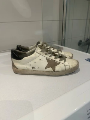  vita sneakers med stjärna -  sneakers i vitt läder med beige mockadetaljer och en brun stjärna på sidan. Svart hälparti och vita snören. Klassisk låg modell med slitna detaljer för en cool och avslappnad look. Pris kan diskuteras och skickas inom en dag