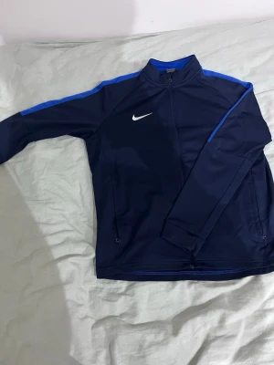 Mörkblå träningsjacka från Nike - Säljer en mörkblå träningsjacka från Nike med vita logotypen på bröstet och blå detaljer längs axlarna. Jackan har dragkedja framtill, ståkrage och två sidofickor med dragkedja. Perfekt för sport och träning.