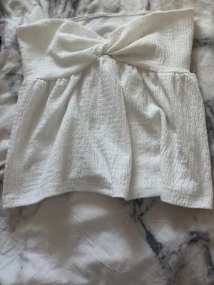 Vit bandeau tubtopp med rosett - Söt vit bandeau tubtopp med rosettdetalj framtill. Toppen är axelbandslös och har en lätt rynkad struktur i tyget som ger en luftig känsla. Perfekt för varma dagar och enkel att styla med olika accessoarer.