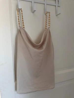 Champagnefärgad topp med kedja - Champagnefärgad linnetopp med breda guldfärgade kedjor som axelband. Toppen har en draperad urringning och ärmlös design. Materialet är glansigt och ger en lyxig känsla.