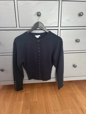 Mörkblå cardigan med knappar - Stilren mörkblå cardigan från Gina Tricot med rund halsringning och silverfärgade knappar längs hela framsidan samt vid ärmsluten. Tröjan har en smal passform och är tillverkad i ett mjukt, tunt material som passar perfekt till lager på lager.