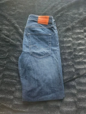 Blå jeans - Säljer ett par blå jeans med klassisk femficksdesign och rak passform. Jeansen har en snygg ljusblå tvätt och ett brunt läderpatch på linningen. Materialet är denim, vilket ger en skön och slitstark känsla. Perfekta för dig som gillar tidlös stil.