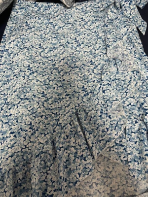 Blå blommig omlottkjol med rosett - Säljer en blå kort omlottkjol med blommigt mönster och en fin rosett i sidan. Kjolen har volangdetaljer och är tillverkad i ett lätt och luftigt material som passar perfekt för varma dagar. Perfekt för dig som gillar romantiska och trendiga plagg.
