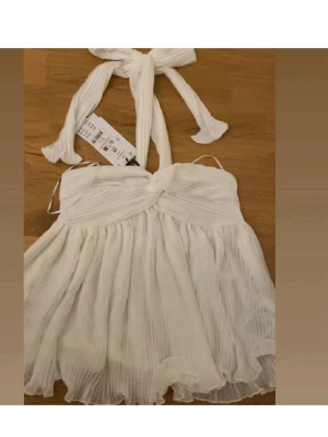 Vit omlott linnetopp med knyt - Säljer en vit linnetopp från H&M med omlott-design och knytband i nacken. Toppen har plisserad, flowy nederdel och är ärmlös med axelbandslös stil. Perfekt för dig som gillar ljusa och luftiga plagg.