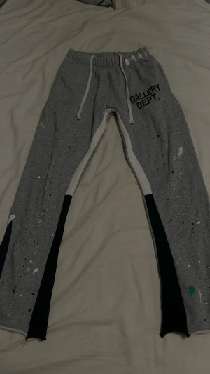 Grå mjukisbyxor från Gallery Dept. - Snygga grå mjukisbyxor från Gallery Dept. med vita snören i midjan och coola färgstänk och tryck på benen. Byxorna har elastisk midja och kontrastpaneler längs benen. Perfekt för en avslappnad och trendig look.