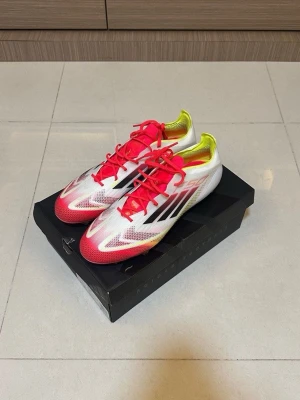 Adidas F50 fotbollsskor vit/röd elite  - Säljer ett par Adidas F50 fotbollsskor med en snygg design i vitt, rött och neon-gult. Skorna har svarta klassiska Adidas-stripes på sidan, röda snören och en lätt, smidig form. Perfekta för snabba spelare som vill ha grymt grepp och stil på planen.