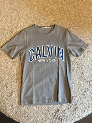 Grå t-shirt från Calvin New York - Snygg grå t-shirt med stort tryck 'CALVIN NEW YORK' i blått och vitt på bröstet. Klassisk rund hals och korta ärmar. Perfekt för dig som gillar enkel och stilren design med en touch av streetwear.