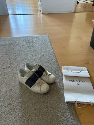 Valentino vita sneakers med svart detalj - Snygga vita sneakers från Valentino med bred svart läderdetalj över snörningen. Klassisk låg modell med rund tå och platt sula. Perfekta för dig som vill ha en stilren och exklusiv look. Skorna är tillverkade i läder och har Valentino-logga på sulan. Man får med påsen och boxen om man vill för den är ganska sönder.
