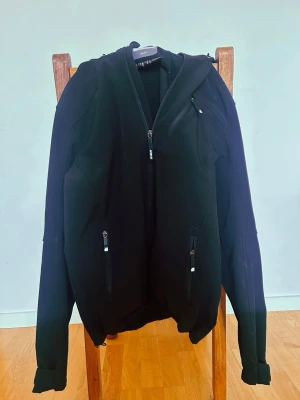 Svart softshelljacka från McKinley - Snygg svart softshelljacka från McKinley med huva och flera dragkedjeförsedda fickor. Jackan har en sportig look, justerbara ärmslut och diskreta loggor på ärm och huva. Perfekt för blåsiga dagar och outdoor-aktiviteter.