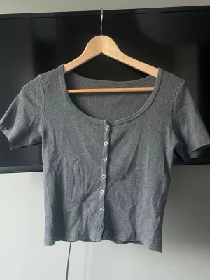 Grå ribbstickad topp med knappar - En grå ribbad topp med korta ärmar och rundad halsringning. Toppen har knappar framtill och är croppad i modellen. Perfekt för att skapa en avslappnad och trendig look. Ärmarna är avklippta men annars så är toppen i ett jättebra skick! Finns ingen lapp men tröjan är köpt från Brandy Melville och storlek S❣️