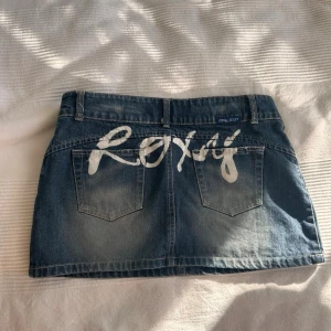 Kort jeanskjol från Roxy - Säljer en cool kort jeanskjol från Roxy med  femficksdesign och snygg tvätt på tyget. Kjolen har en vit logotyp på baksidan och är tillverkad i slitstarkt denim med knapp och dragkedja framtill. Perfekt för dig som gillar streetstyle.