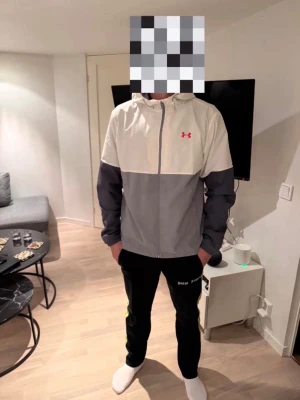 Under Armour vindjacka med huva - Snygg vindjacka från Under Armour med tvåfärgad design i ljusbeige och grått. Jackan har huva, dragkedja framtill och meshfoder. Under Armour-loggan i rosa på bröstet ger en sportig touch. Perfekt för att hålla vinden borta under utomhusaktiviteter.