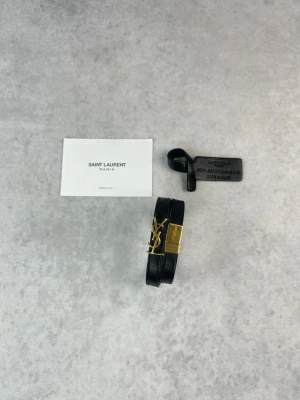 Saint Laurent svart läderarmband - Snyggt svart armband från Saint Laurent i äkta läder med dubbla remmar och ikoniskt YSL-spänne i guldmetall. Perfekt accessoar för dig som gillar stilrena detaljer och exklusiv känsla. Justerbar storlek och klassisk design.