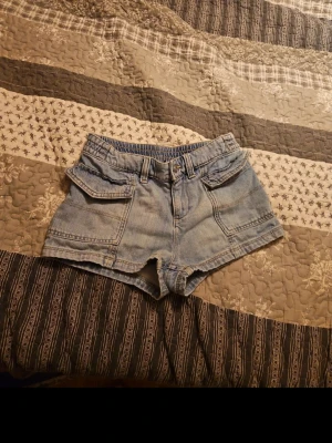 Blå denimshorts med fickor - Säljer ett par klassiska blå denimshorts med stora framfickor och bälteshällor. Shortsen har en avslappnad passform och är tillverkade i bomull. Perfekta för varma dagar och enkel att matcha med olika toppar.