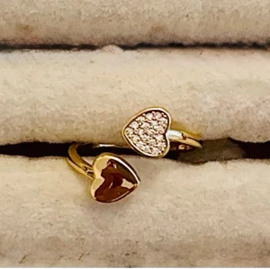 Guldfärgad ring med hjärtan - Snygg guldfärgad ring med två hjärtformade detaljer, varav den ena är täckt med gnistrande stenar och den andra har en blank yta. Perfekt för dig som gillar romantiska accessoarer och vill ha något som sticker ut.