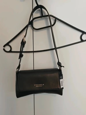 Brun läderhandväska Stockholm Design Group nypris ca 1000 kr - Nypris ca 1000 kr. Stilren svart handväska i läder från Stockholm Design Group med justerbar axelrem och guldfärgad logotyp på framsidan. Väskan har en klassisk rektangulär form och praktiskt lock. Perfekt för dig som gillar minimalistisk design.