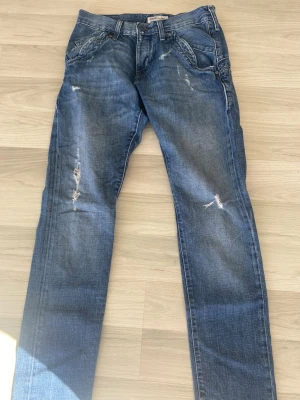 Blå slitna jeans från Lee - Säljer ett par blå jeans från Lee med slitningar och distressed detaljer på benen. Klassisk femficksmodell med raka ben och normal midja. Jeansen är gjorda i bomull och har en avslappnad streetstil. I storlek w26
