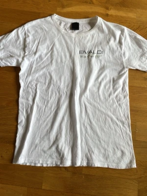 Vit BVALDI t-shirt med tryck - Vit t-shirt från BVALDI med tryck på bröstet och stort tryck på ryggen med två tennisrack och texten 'Bistro Tennis Club'. Klassisk passform och rund hals. Perfekt för dig som gillar sportig stil.