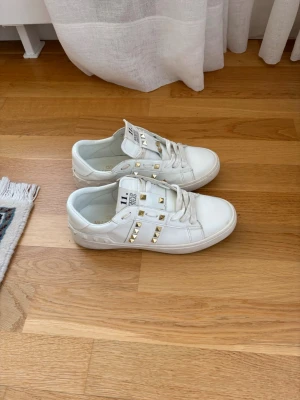 Vita sneakers med guldnitar från Valentino - Snygga vita sneakers från Valentino med coola guldnitar på sidorna och tungan. Skorna har klassisk låg profil, vita skosnören. Perfekt för dig som vill sticka ut med en exklusiv och trendig look. Osäker på varans äkthet då jag köpt dem på nätet utan orginalkvitto.