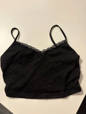 Svart spetsdetaljerad crop top oanvänd  - Oanvänd Svart crop top från SHEIN med tunna axelband och spetsdetaljer längs v-ringad hals. Toppen är tillverkad i mjukt tyg och har en enkel, stilren design som passar perfekt till jeans eller kjol. Strl M