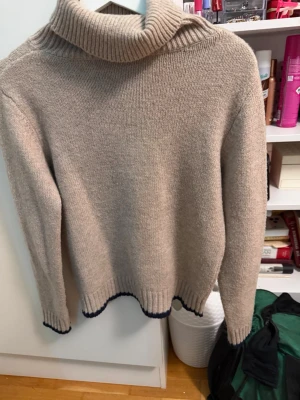 Beige polotröja med blå detaljer - Stickad beige polotröja med ribbad krage och blå kontrastdetaljer vid ärmslut och nederkant. Tröjan har en avslappnad passform och långa ärmar, perfekt för kyliga dagar. Materialet ser ut att vara mjukt och skönt, troligen ull eller ullmix.