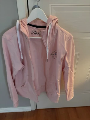 Ljusrosa hoodie från Polo Club - Ljusrosa hoodie med dragkedja från Polo Club Saint Vincent. Tröjan har huva med vita snören, två fickor framtill och diskret logga på bröstet. Perfekt för en avslappnad och cool look. Materialet är mjukt och bekvämt, troligen bomull.