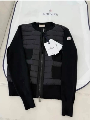 Svart cardiganjacka från Moncler - Snygg svart cardiganjacka från Moncler med quiltad front och stickade ärmar. Jackan har dragkedja, två fickor fram och Moncler-logga på ärmen. Materialmix med vadderad front och mjuka stickade partier ger en modern look.