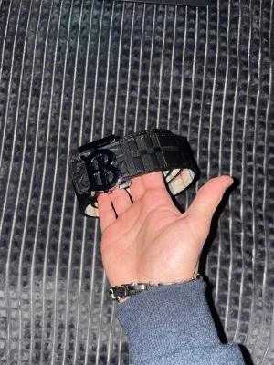 Svart Burberry bälte i skinn - Säljer ett svart Burberry bälte i skinn med rutigt mönster och stor BB-logga i metall på spännet. Bältet har även ett präglat Burberry-riddare motiv. Perfekt accessoar för att lyfta din outfit.