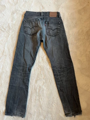 Grå Levi's raka jeans - Säljer ett par grå raka jeans från Levi's med klassiska fem fickor och läderpatch bak i midjan. Jeansen har en snygg tvättad look och råa kanter vid bensluten. Perfekta för en avslappnad stil. W28 L30