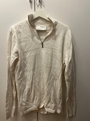 Vit stickad tröja med dragkedja Selected - Vit stickad tröja från Selected Homme med halv dragkedja vid halsen. Tröjan har strukturerade partier i ett diskret rutmönster och är långärmad. Perfekt för lager-på-lager och enkel att matcha med andra plagg.