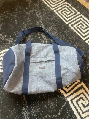 Blå weekendbag från Hollister - Snygg och rymlig weekendbag från Hollister i blått tyg med mörkblå detaljer. Väskan har både axelrem och handtag samt en praktisk ytterficka. Perfekt för resor eller gymmet, med stilren design och diskret logga framtill.