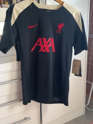 Liverpool tränings t-shirt Nike svart M - Snygg svart tränings t-shirt från Nike x Liverpool FC med röda detaljer och AXA-logga på bröstet. Tröjan har beiga partier på axlarna och är i slim fit-modell. 