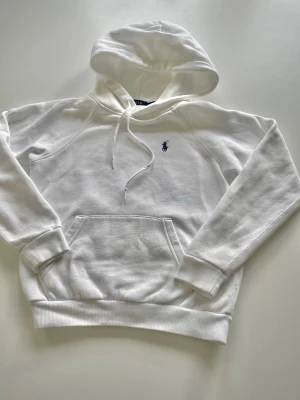 Vit hoodie från Polo Ralph Lauren - Klassisk vit hoodie från Polo Ralph Lauren med broderad logga på bröstet. Hoodien har känguruficka, dragsko i huvan och ribbade muddar. Perfekt för en clean och stilren look.