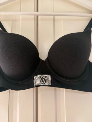 Svart push-up bh från Victoria's Secret - Snygg svart push-up bh från Victoria's Secret med vadderade kupor och justerbara axelband. Framtill finns en glittrig detalj med VS-logga i strass. Perfekt för dig som vill ha extra lyft och stilren look.