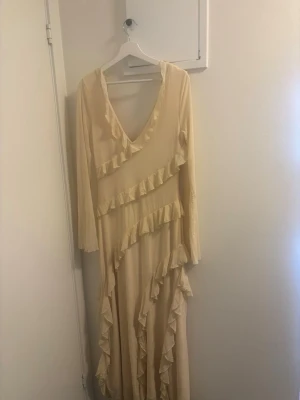 Långklänning med volang och v-ringning - Elegant ljusbeige långklänning med v-ringning och långa ärmar. Klänningen har omlottkänsla och dekorativa volanger som löper diagonalt över hela plagget. Perfekt för dig som gillar en romantisk och bohemisk stil. Ordinarie pris 450