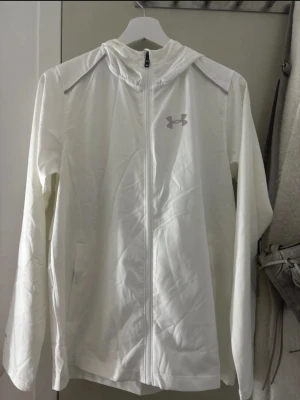 Vit vindjacka från Under Armour - Snygg vit vindjacka från Under Armour med dragkedja framtill och huva. Jackan har diskret logga på bröstet och är tillverkad i lätt, vindtätt material. Perfekt för blåsiga dagar och sportiga outfits.