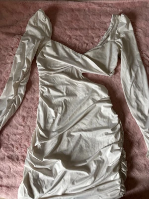 Vit långärmad kortklänning - Trendig vit kortklänning med långa puffärmar och snygg v-ringning. Klänningen har draperade detaljer längs sidorna och en lätt omlott-effekt vid bysten. Perfekt för dig som gillar en stilren och modern look.