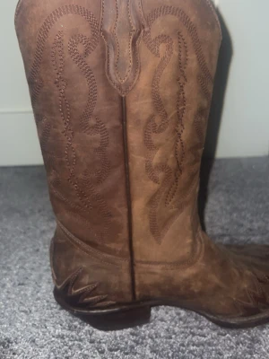 Bruna cowboy boots med broderi - Snygga bruna cowboy boots i läder med klassiskt broderat mönster längs skaftet och spetsig tå. Bootsens klack är medelhög och sulan är robust. Perfekta för dig som vill ha en western-inspirerad look.