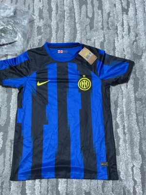 Inter Milan Hemma 23/24 - Säljer en Inter Milan matchtröja från Nike i blått och svart med gula detaljer. Tröjan har klubbens emblem och Nike-logga på bröstet samt klassiska vertikala ränder. Tillverkad i lätt och ventilerande material, perfekt för fotbollsträning.