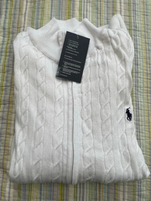 Vit kabelstickad kofta från Polo Ralph Lauren - Vit kabelstickad kofta med dragkedja från Polo Ralph Lauren. Klassisk design med ribbade muddar och broderad marinblå logga på bröstet. Perfekt för en stilren och fräsch look.