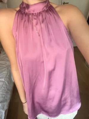 Lila halterneck linnetopp i satin - Snygg lila halterneck linnetopp i glansigt satinmaterial. Toppen är ärmlös och har hög hals med lätt rynk vid halsringningen. Perfekt för dig som vill ha en chic och trendig look.
