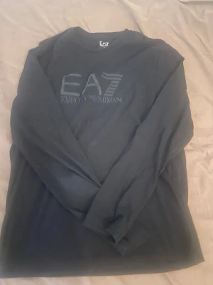 Svart långärmad t-shirt EA7 Armani - Svart långärmad t-shirt från Emporio Armani EA7 med diskret logga framtill. Klassisk rund halsringning och stilren design som passar till det mesta. Tillverkad i mjukt material för en bekväm känsla.