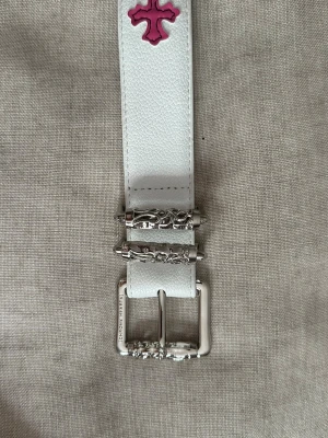 Vitt och färgat roller belt - Unikt vitt bälte i skinn med dekorativa kors i rosa, gult, grönt och blått längs hela bandet. Spännet är i silverfärgad metall med snygga ingraverade detaljer. Perfekt accessoar för att lyfta din outfit med lite färg och edge. 110cm